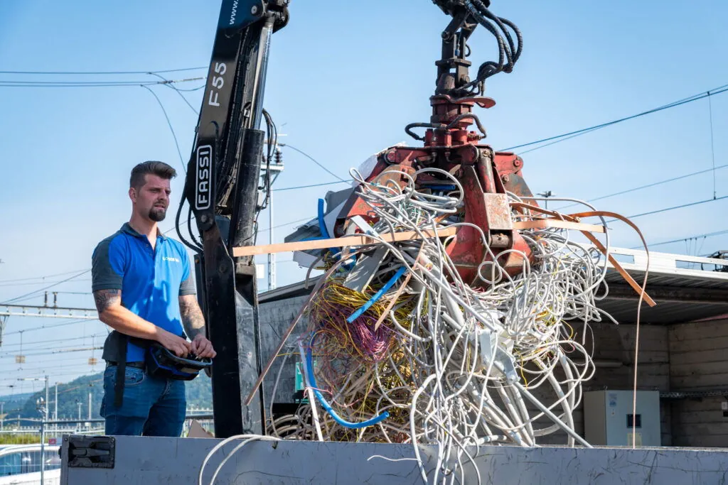 Mitarbeiter steuert per Fernbedienung einen Kran, der Altkabel auf einen Lastwagen lädt